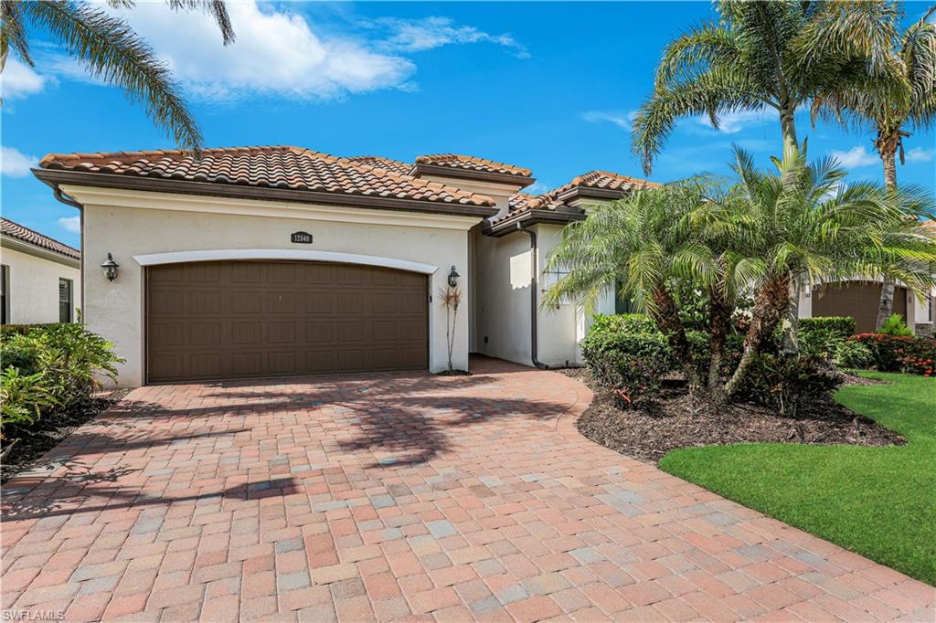 Stucco exterior with a tile roof, featuring a paver driveway, an arched garage door, and mature palm tree landscaping