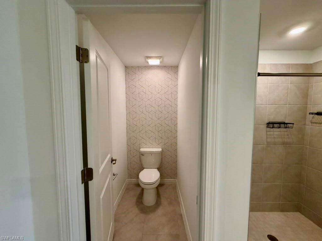 Full bathroom featuring a commode alcove with patterned wall covering, a built-in shower with tile surround, and neutral tile flooring