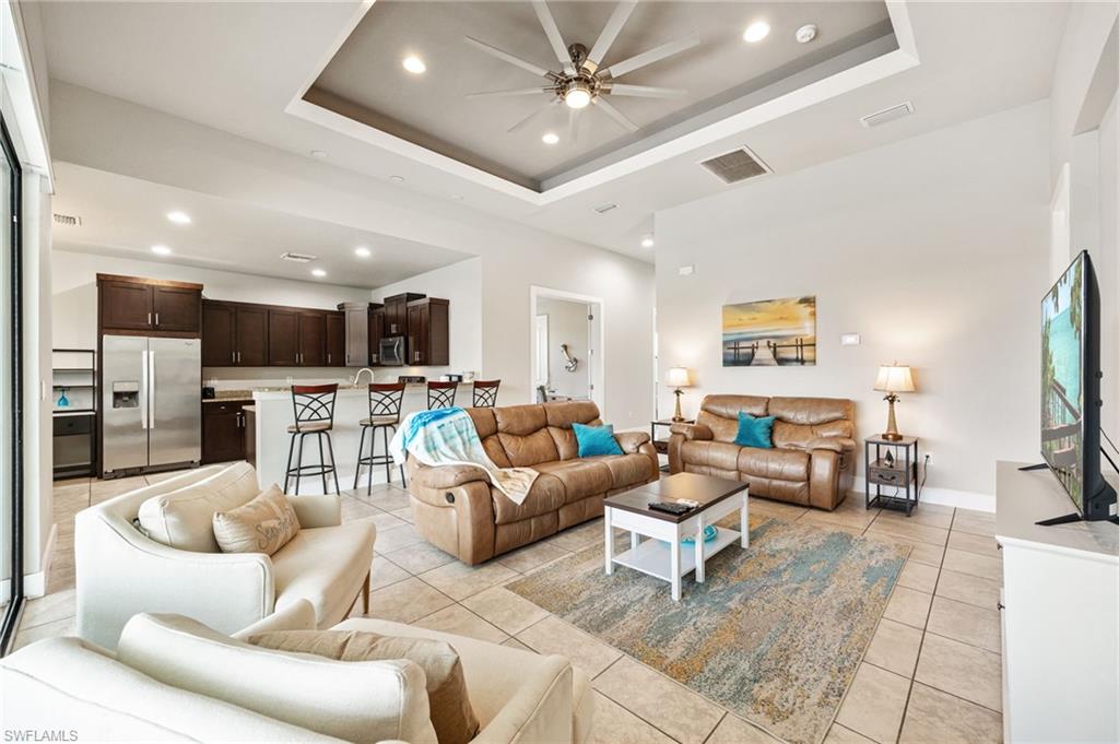 Open-concept living space featuring neutral tile flooring, a tray ceiling with recessed lighting and ceiling fan, and a kitchen with dark wood cabinetry and stainless steel appliances