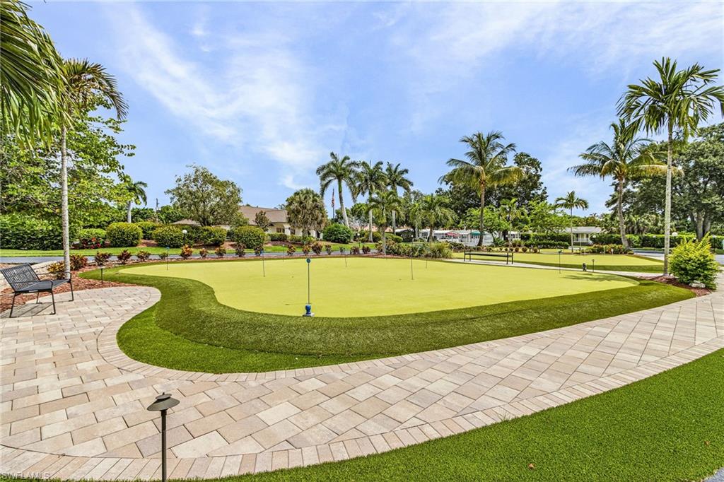 Expansive putting green with multiple flag positions, surrounded by a paver walkway, artificial turf, and mature palm trees.