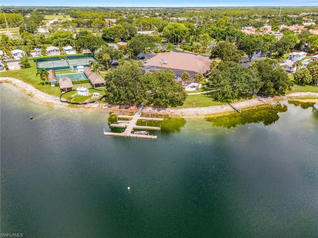 Aerial lake view featuring community boat dock with multiple slips, a large clubhouse, two tennis courts, and mature trees throughout the grounds.
