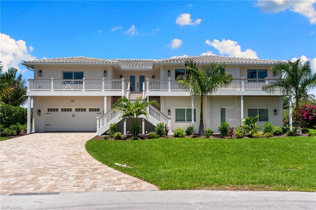 Contemporary elevated residence featuring a two-car garage, upper-level balcony, dual exterior staircases, and a paver driveway
