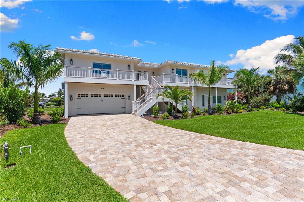 Elevated coastal residence featuring a paver driveway, lush green lawn, mature palm trees, multiple balconies with white railings, and an integrated two-car garage