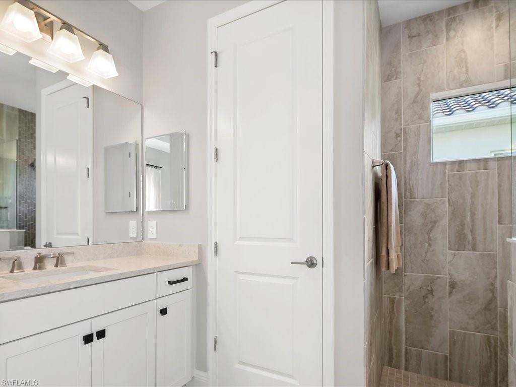 Bathroom featuring a double vanity with light-toned countertop, white cabinetry with dark hardware, a shower with floor-to-ceiling tile, and a frosted transom window