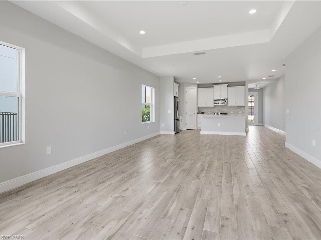 Spacious open-concept living area featuring light wood-finish flooring, a contemporary kitchen with white cabinetry, stainless steel appliances, and a tiled backsplash