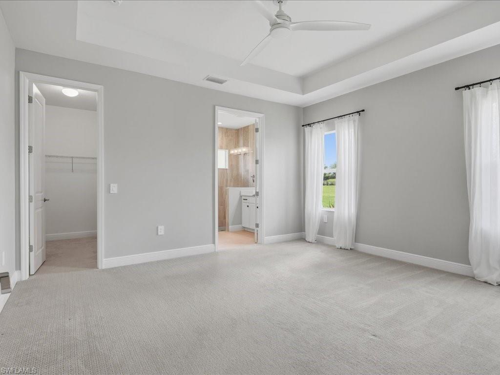 Spacious room featuring light gray carpeting, a tray ceiling with a modern ceiling fan, and multiple windows with white sheers