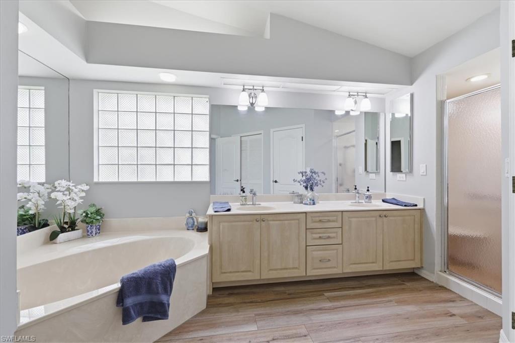 Bathroom featuring a large soaking tub, glass block window, double vanity with light wood cabinetry, a frameless shower, and wood-finish flooring