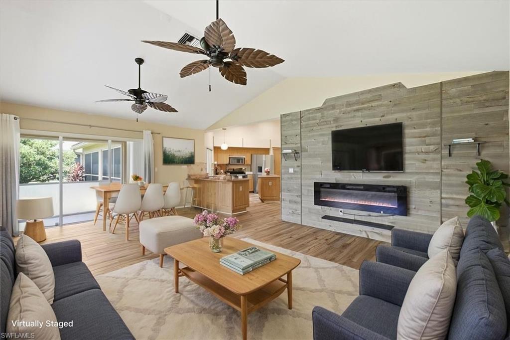 *VIRTUALLY STAGED* Spacious living area featuring vaulted ceilings, wood-finish flooring, a wall-mounted electric fireplace, and a kitchen with light wood cabinetry and a breakfast bar