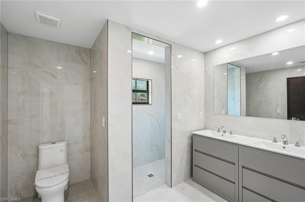 Contemporary bathroom featuring a dual vanity with undermount sinks and chrome fixtures, a spacious walk-in shower, a toilet, recessed lighting, and large format tiled walls