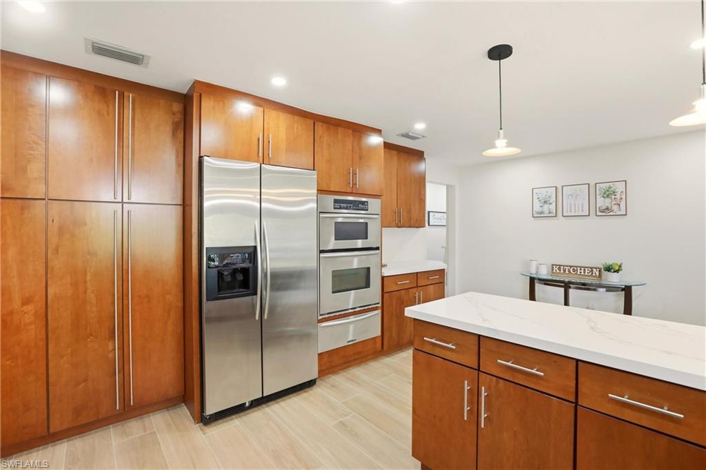 Kitchen featuring brown cabinetry, appliances with stainless steel finishes, decorative light fixtures, light stone counters, and a warming drawer