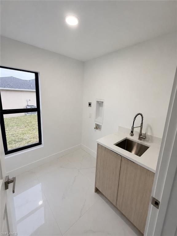 Laundry room featuring a single basin stainless steel utility sink with a pull-down faucet, light wood-finish cabinetry, white countertop, and white large-format tile flooring