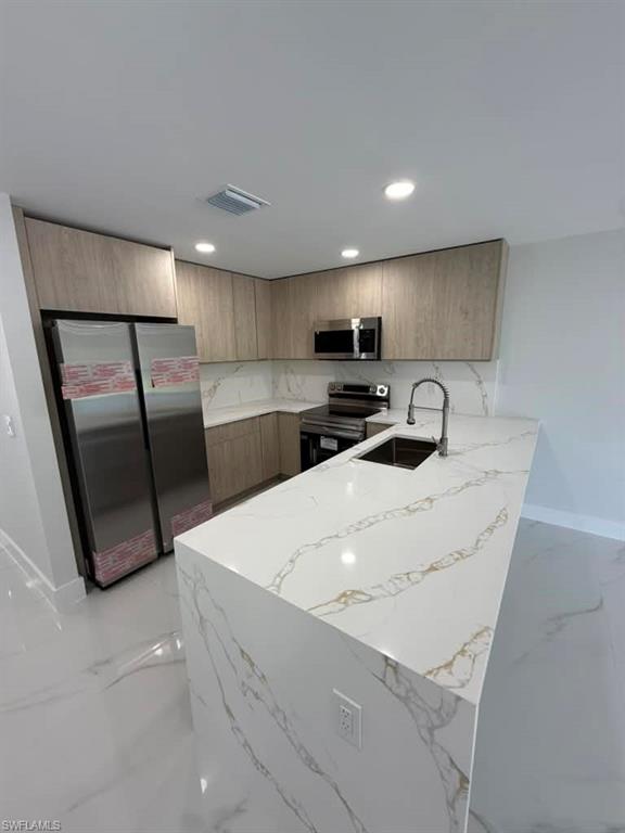Contemporary kitchen featuring a spacious island with a waterfall edge, stone countertops with gold veining, stainless steel appliances, light wood-finish cabinetry, and recessed lighting