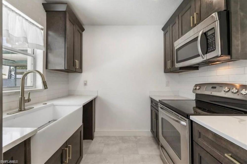 Galley kitchen featuring dark wood-finish cabinetry, white countertops, a white farmhouse sink with a brushed nickel faucet, and stainless steel appliances including a range and microwave