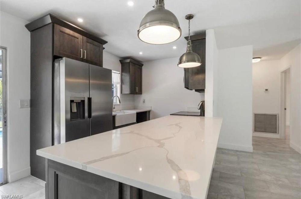 Kitchen featuring a white countertop island with gray veining, dark wood-finish cabinetry, stainless steel refrigerator, farmhouse sink, and two pendant light fixtures
