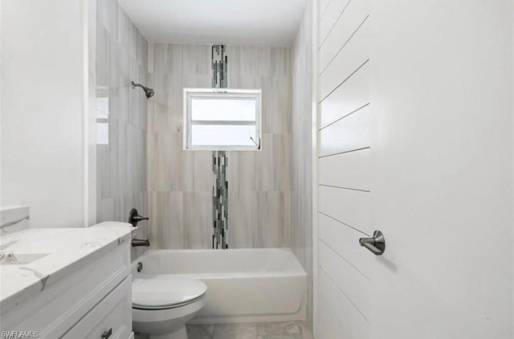 Bathroom featuring a built-in tub with a shower fixture, light-toned vertical wall tiling with a mosaic accent, a small window, a white vanity with a marble-finish countertop, and a toilet