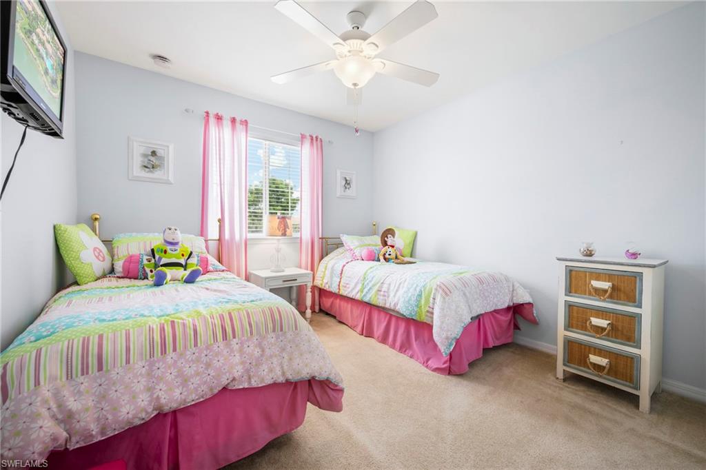 Bedroom featuring light blue walls, window with blinds and pink sheer curtains, beige carpet flooring, and a ceiling fan with integrated lighting