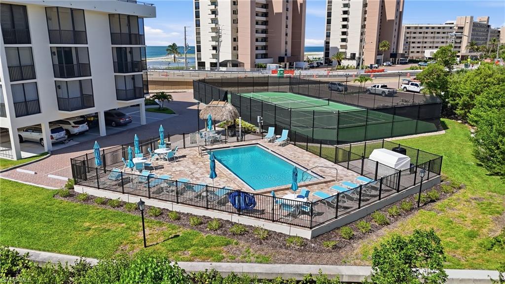 Outdoor swimming pool area with paver decking, black metal perimeter fencing, and a nearby tennis court - Virtually Edited Image