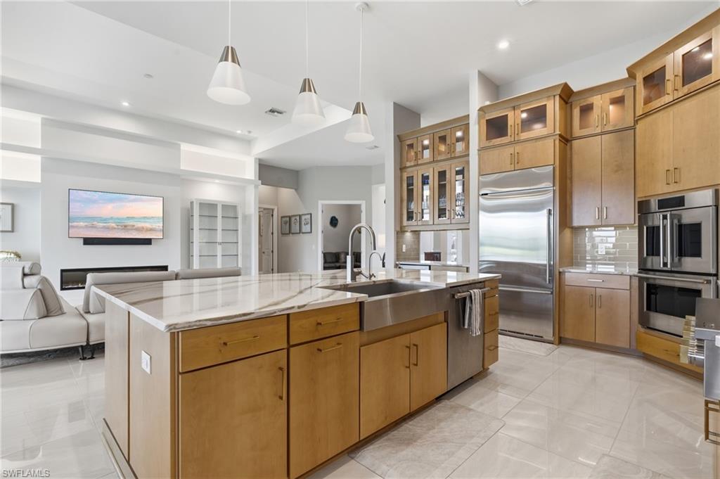 Spacious kitchen featuring a large central island with a farmhouse-style sink, light wood cabinetry, stainless steel appliances, and a tile backsplash