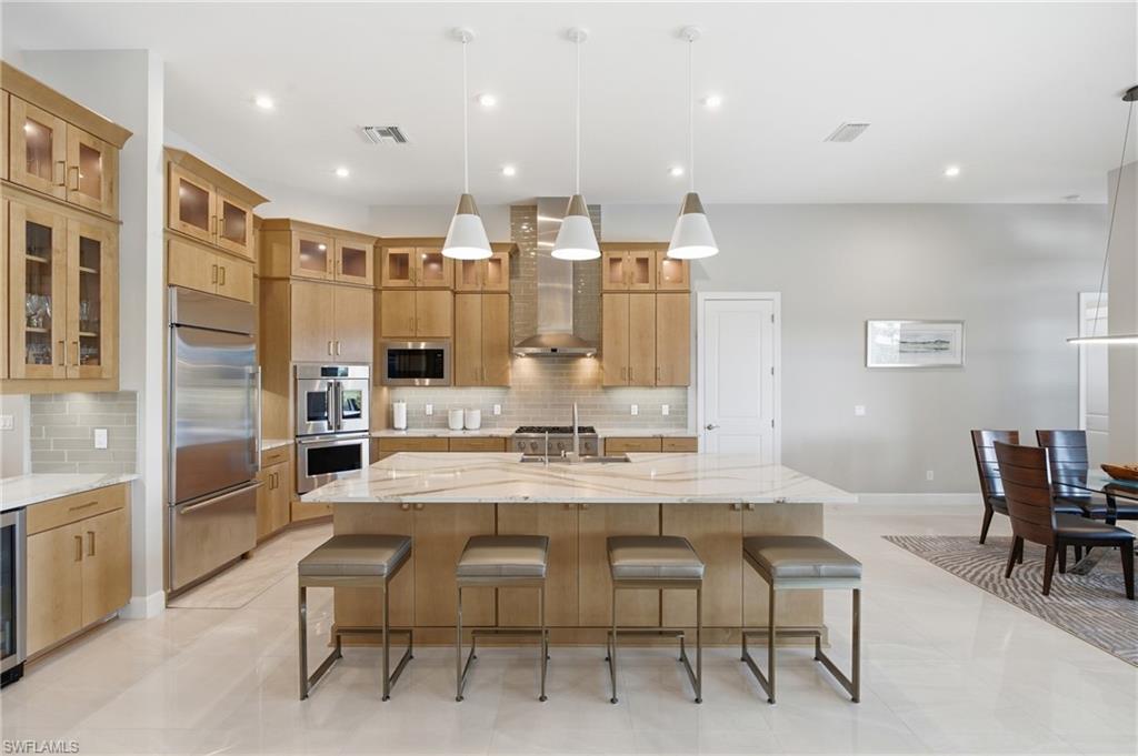 Modern kitchen featuring an expansive center island with seating, light wood-finish cabinetry, stainless steel appliances, and a tiled backsplash