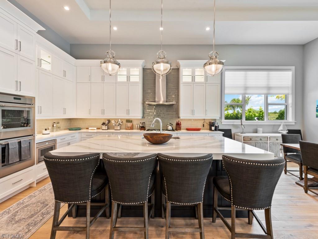 Kitchen with backsplash, light stone counters, double oven, light hardwood / wood-style floors, and wall chimney exhaust hood