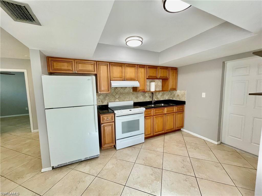 Kitchen featuring natural wood cabinetry, black countertops, tile backsplash, tile flooring, and white appliances including a refrigerator and range