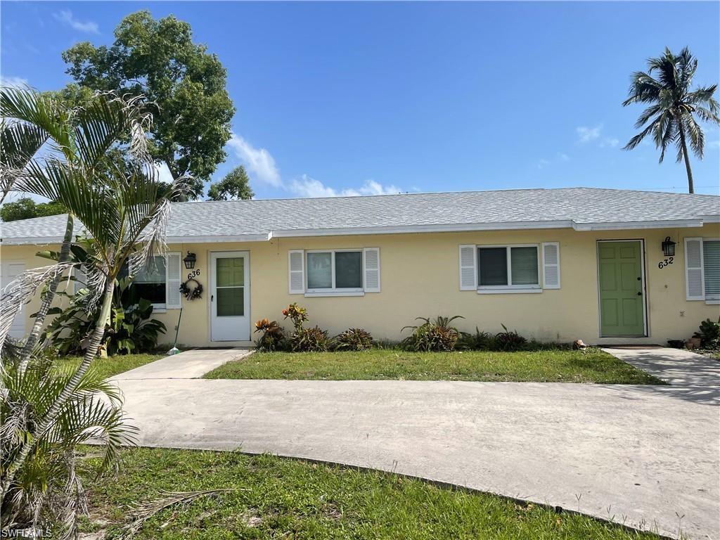 Exterior featuring a light yellow facade, white window shutters, a light gray shingled roof, a paved driveway, and mature landscaping