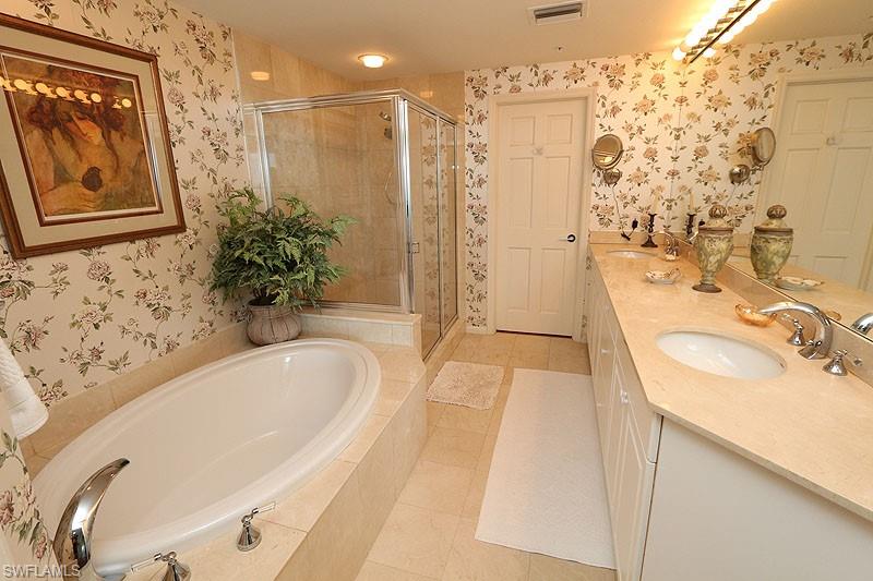 Bathroom featuring an oval soaking tub with deck-mounted fixtures, a glass-enclosed shower, dual undermount sinks set in a light-toned countertop, and light-toned floor tiling