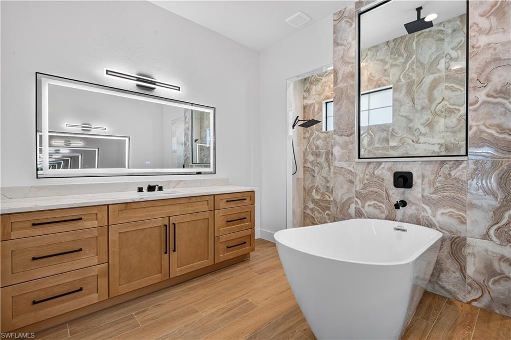 Bathroom featuring a freestanding soaking tub, large vanity with light wood-finish cabinetry, stone-look tile accents, and modern black fixtures