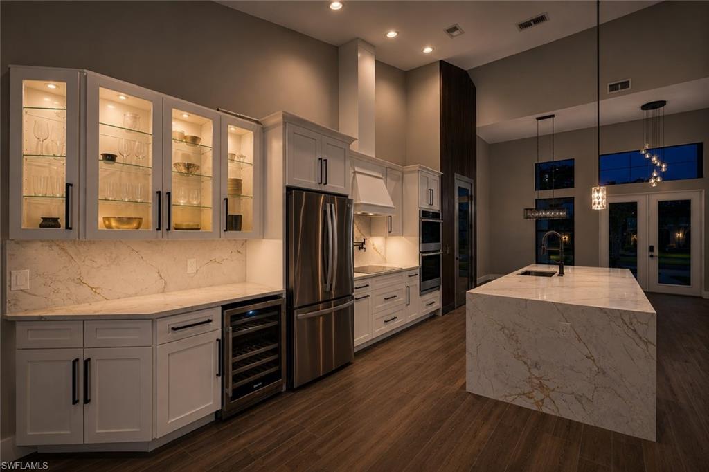 Contemporary kitchen featuring white cabinetry, integrated stainless steel appliances, a waterfall island with a sink, wood-finish flooring, and high ceilings with recessed lighting