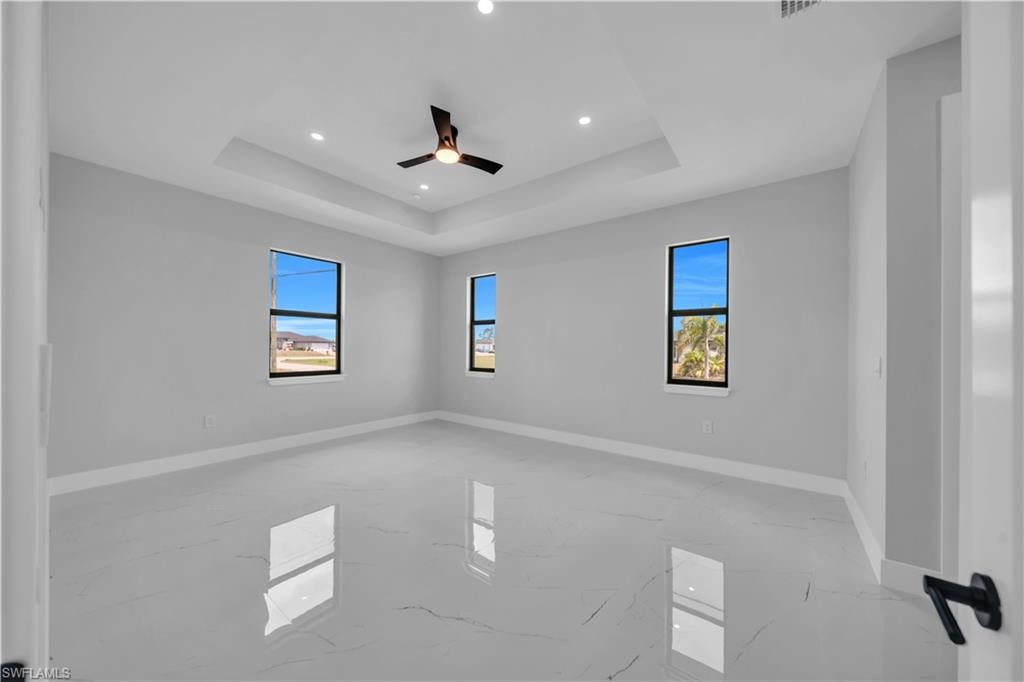 Spacious room featuring tray ceiling, recessed lighting, a ceiling fan, three windows, and large-format tile flooring