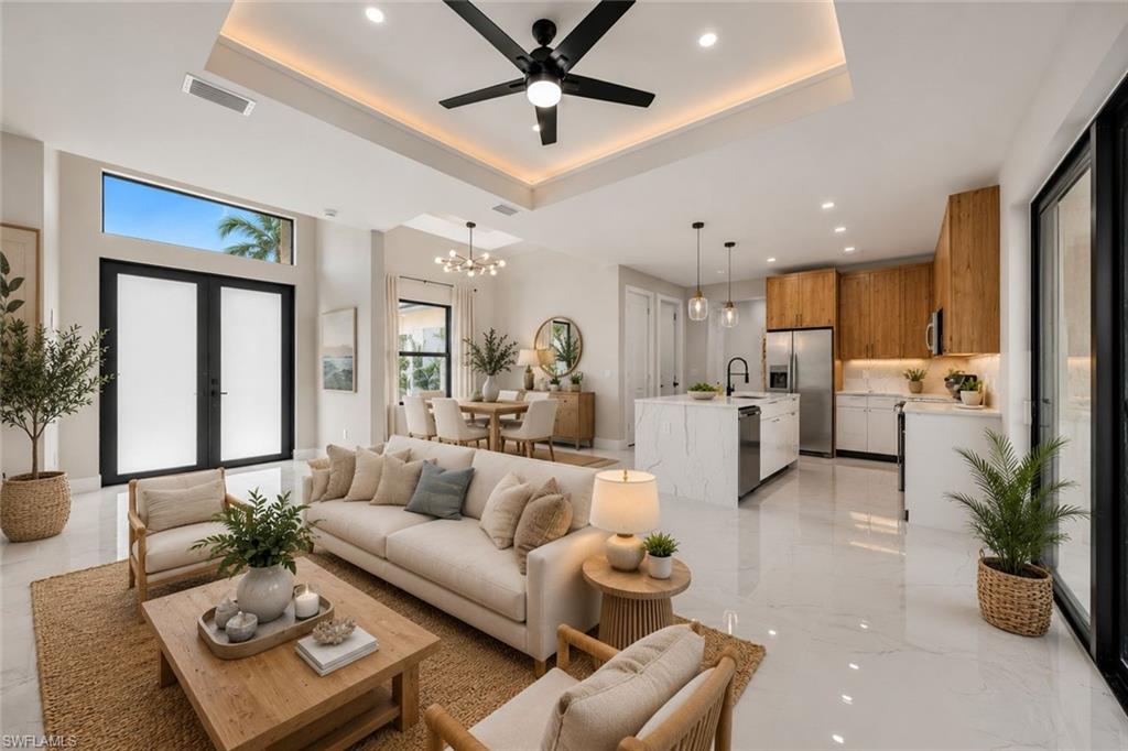 Open-concept living space featuring a tray ceiling with recessed lighting and a ceiling fan, large format tile flooring, a kitchen with wood cabinetry and a waterfall-edge island, and a dining area with a modern chandelier