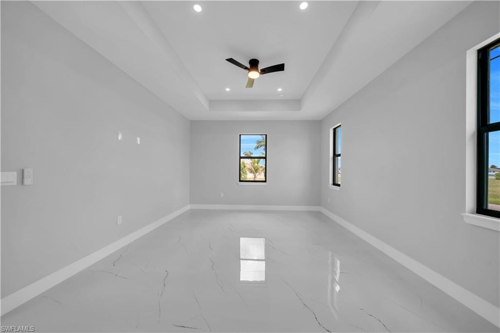Spacious room featuring tray ceilings with recessed lighting, a ceiling fan, multiple windows with black framing, and large-format porcelain tile flooring