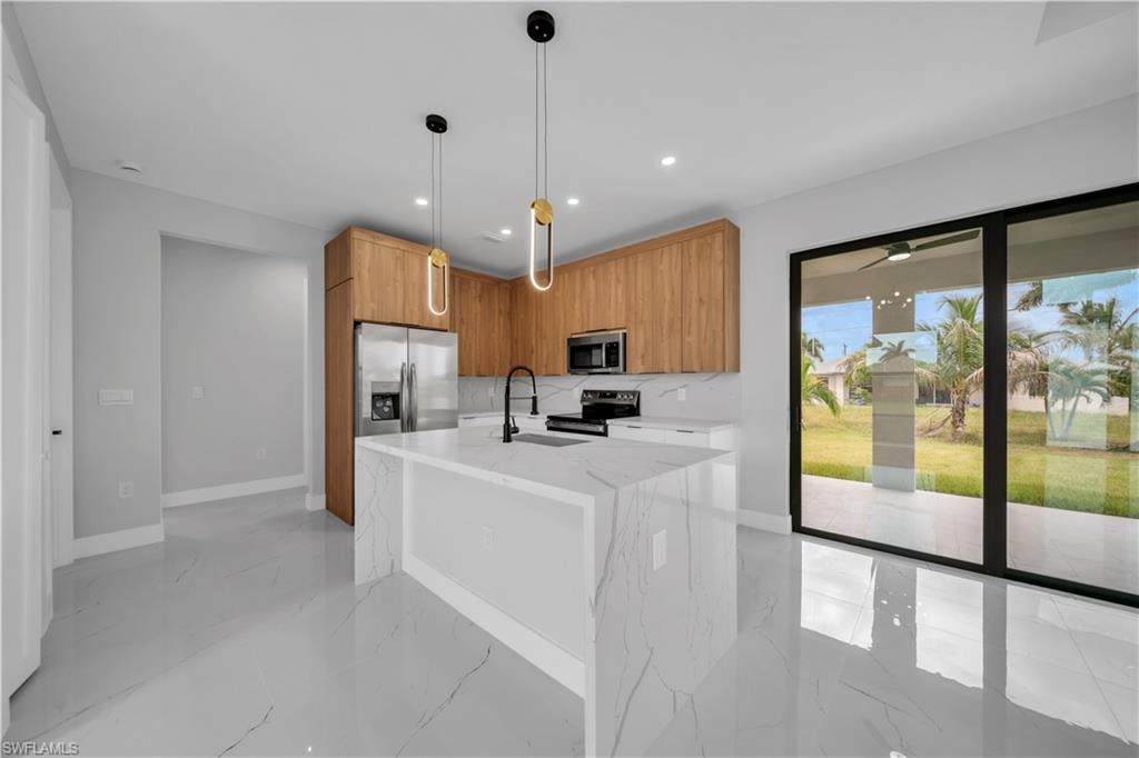 Kitchen featuring an oversized island with waterfall edge, wood-finish cabinetry, stainless steel appliances, and sleek pendant lighting