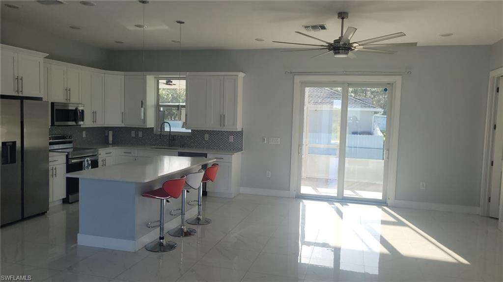Open-concept kitchen and living area featuring white cabinetry, a central island with a light-colored countertop, stainless steel appliances, and a mosaic tile backsplash
