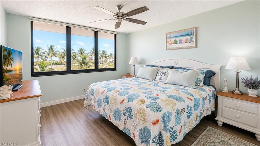 Expansive windows showcasing palm trees, wood-finish flooring, a ceiling fan, a white headboard, and light-toned walls
