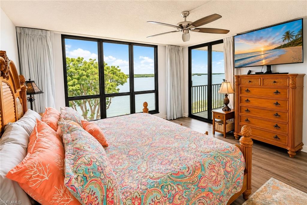 Spacious bedroom featuring large windows, sliding glass door balcony access, water scenery, wood-finish flooring, and a ceiling fan with light fixture