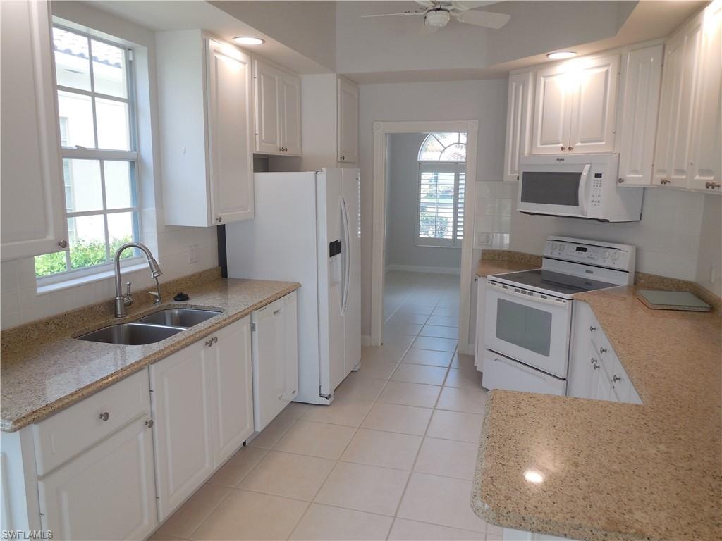 Kitchen featuring white cabinetry, light-toned countertops, a dual basin stainless steel sink with a gooseneck faucet, white appliances, and tiled flooring