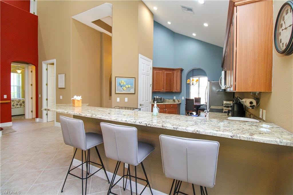 Kitchen featuring light tile patterned flooring, high vaulted ceiling, a breakfast bar area, light stone counters, and kitchen peninsula