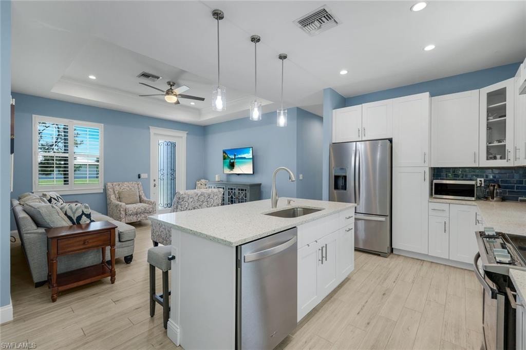 Open-concept kitchen and living area featuring a central island with sink and stainless steel dishwasher, white cabinetry, light countertops, and a blue tile backsplash