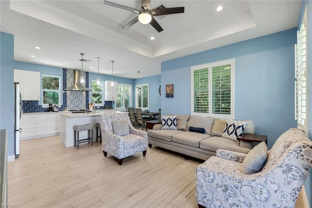 Open-concept living space featuring wood-finish flooring, a tray ceiling with a ceiling fan, and multiple windows with plantation shutters