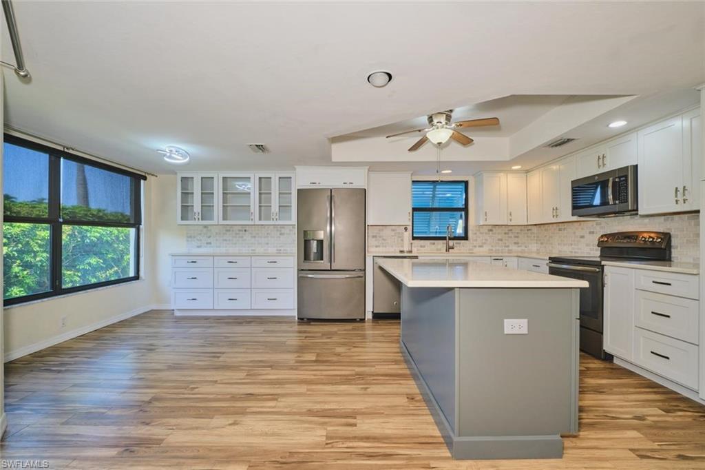 Modern kitchen featuring wood-finish flooring, white cabinetry, a central island with a light-toned countertop, stainless steel appliances, and a tiled backsplash