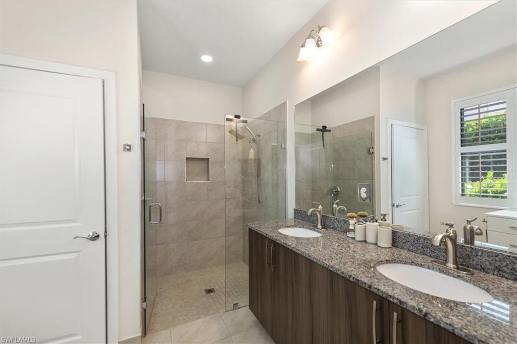 Bathroom featuring a dual vanity with granite countertops and undermount sinks, a large frameless mirror, a glass-enclosed shower with tiled walls and an integrated niche, and a window with plantation shutters