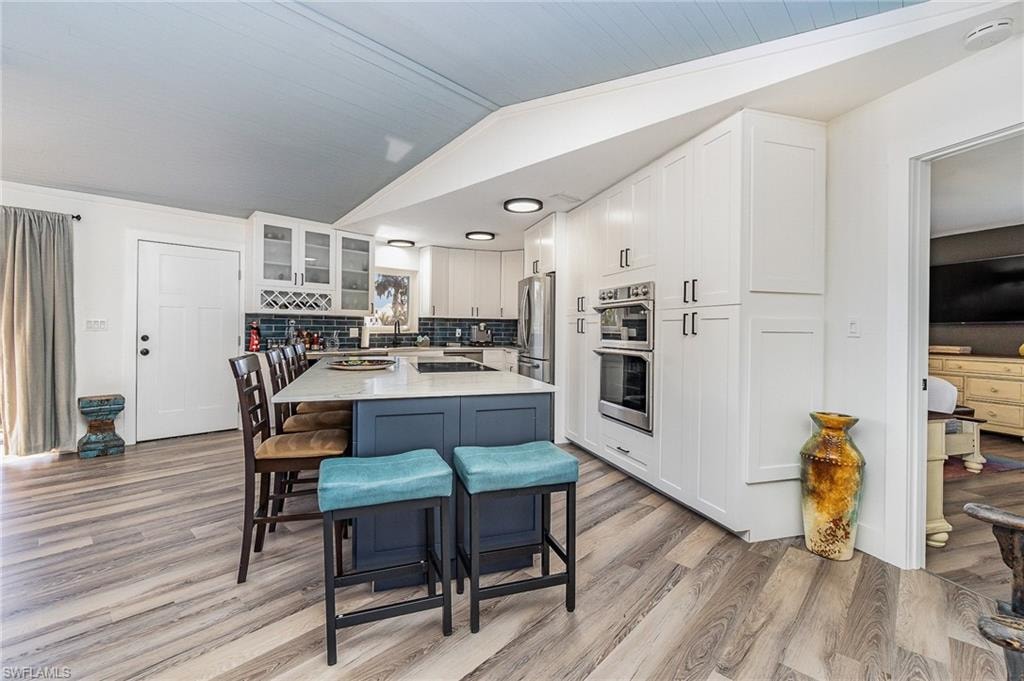 Kitchen featuring a large central island with seating, white shaker cabinetry, stainless steel double ovens, and a dark blue subway tile backsplash