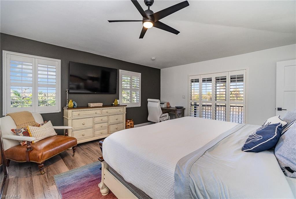 Bedroom featuring wood-finish flooring, a ceiling fan with integrated lighting, white plantation shutters on windows and a sliding door, and a built-in closet door with black hardware