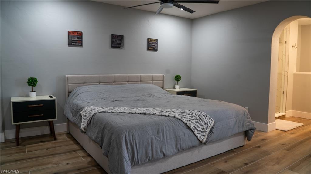 Bedroom featuring wood-finish flooring, a contemporary ceiling fan with integrated lighting, and an arched doorway leading to a walk-in shower with clear glass enclosure
