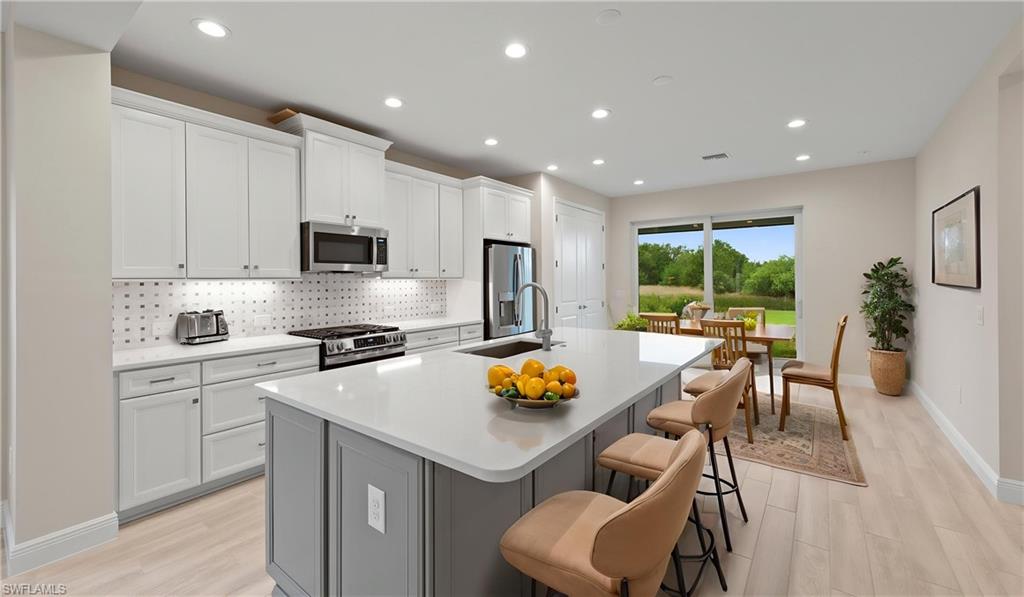A Stone Gray center island topped with Ethereal White quartz and breakfast-bar seating anchors the kitchen, complemented by white Wentworth cabinetry, a GE stainless steel appliance suite with gas range, a stone-tile backsplash, and under-cabinet LED lighting.
