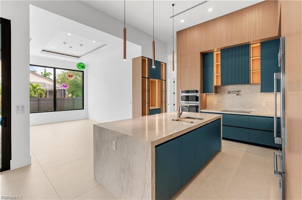 Modern kitchen featuring a large waterfall island with integrated sink, teal cabinetry, wood-panel accents, built-in stainless steel appliances, and light-toned tile flooring