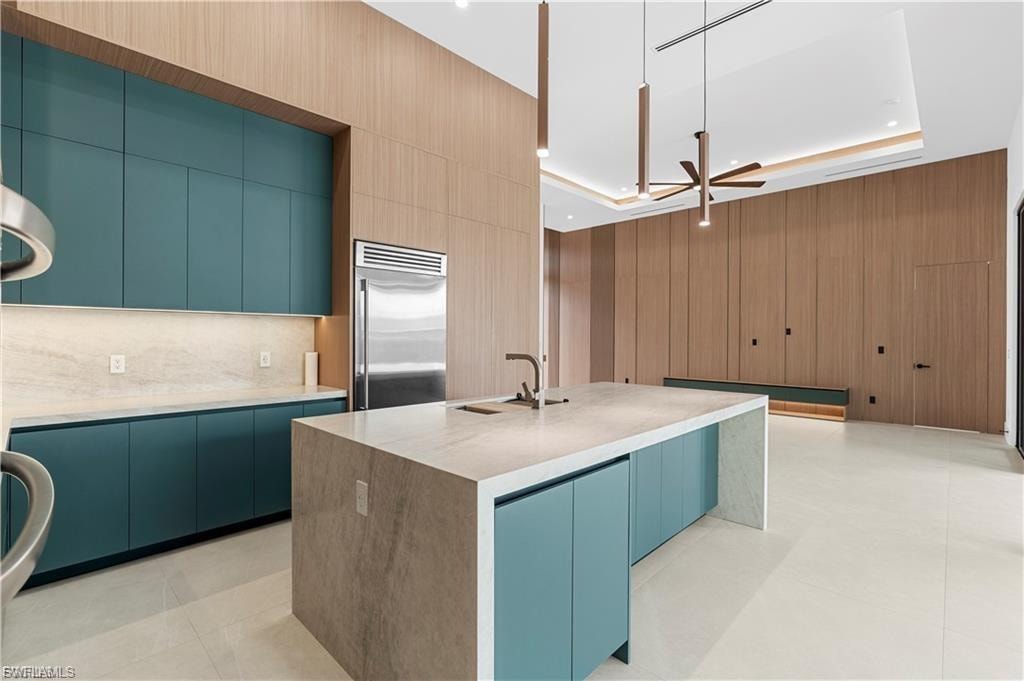 Gourmet kitchen featuring a waterfall island with integrated sink, stainless steel refrigerator, and teal flat-panel cabinetry