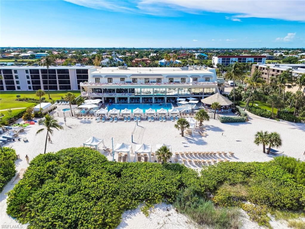 Expansive beachfront property featuring a multi-story building with large windows, a swimming pool with surrounding lounge chairs and cabanas, a thatched-roof structure, and mature palm trees