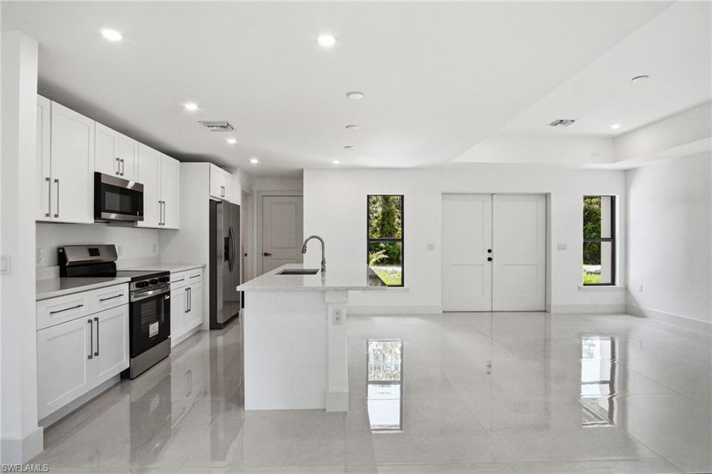 Open-concept kitchen and living area featuring sleek white cabinetry, stainless steel appliances, a center island with an integrated sink, recessed lighting, and large format tile flooring